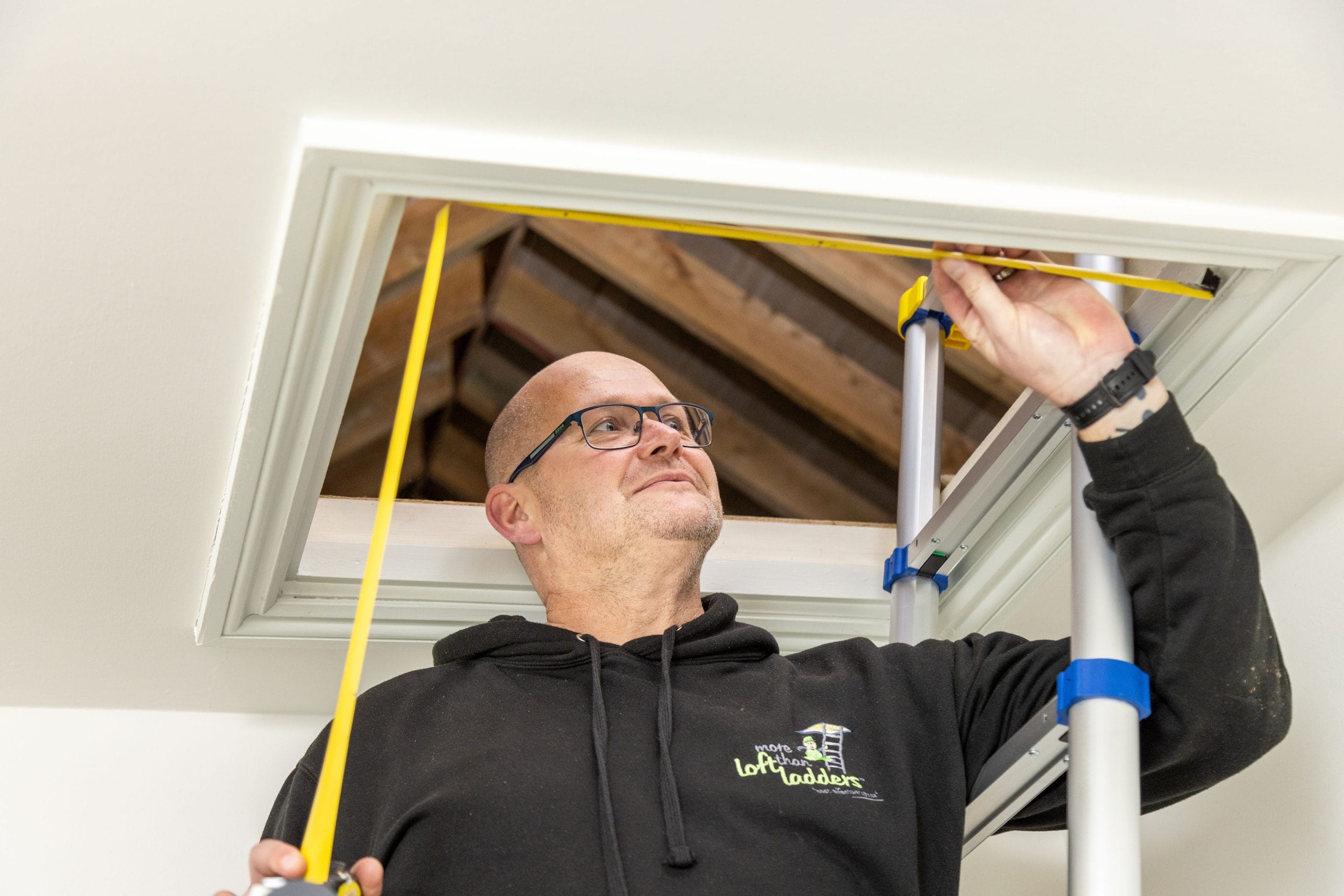 More Than Loft Ladders installer measuring loft hatch opening with tape measure on ladder